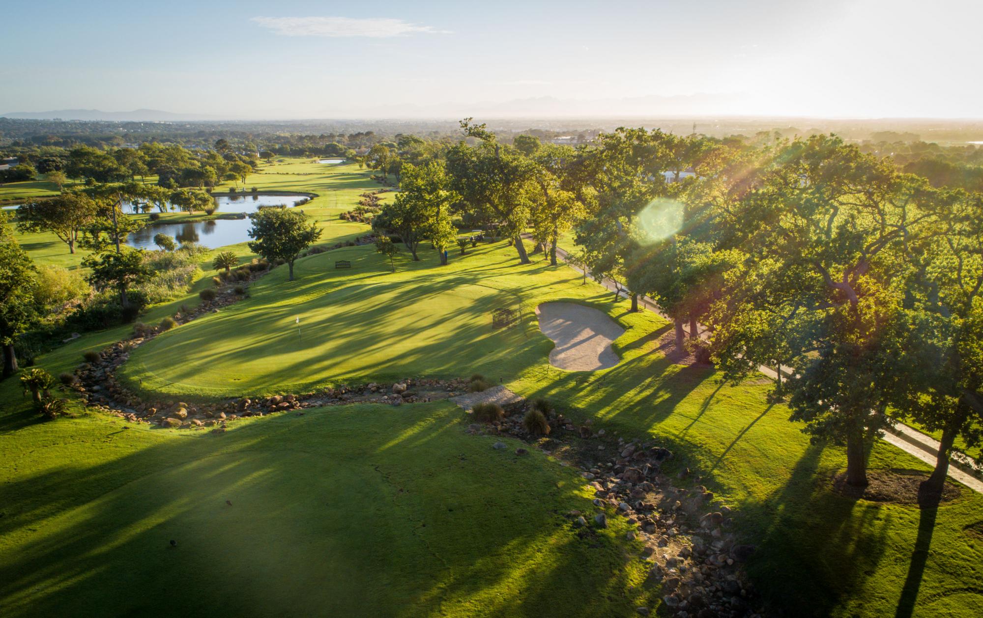 Aerial view over a green and bunker at Steenberg Golf Club in golden late-afternoon light with the Cape mountains beyond