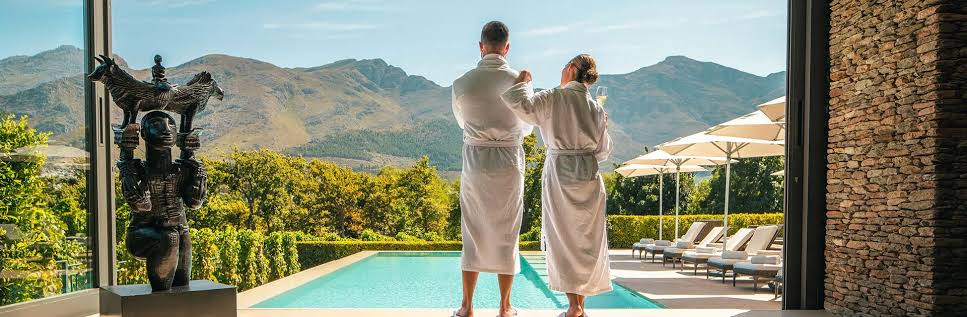 Couple in robes with wine beside a pool at a Winelands hotel with the mountains beyond